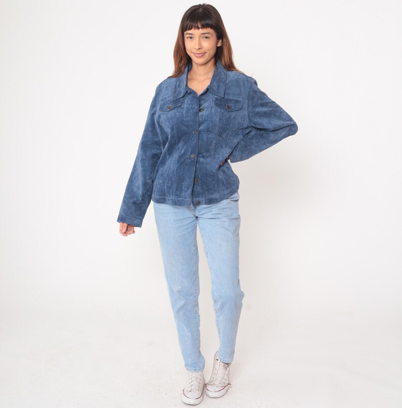 May include: A blue corduroy jacket with button closures and two chest pockets. The jacket is paired with light blue jeans and white sneakers. The outfit is worn by a person with brown hair.
