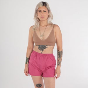 May include: A woman wearing a tan bralette and pink athletic shorts. The shorts have an elasticated waistband and a relaxed fit. The woman is also wearing a necklace.