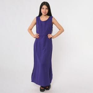 May include: A sleeveless, floor-length, purple dress. The dress features a scoop neckline, a fitted waist, and vertical pleats on the bodice. The skirt has a slight flare and side slits. The model is wearing black platform sandals.