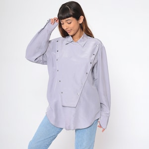 May include: A light purple button-down shirt with long sleeves and a unique asymmetrical design. The shirt features silver-colored buttons and is paired with light blue jeans. The model is wearing the shirt.