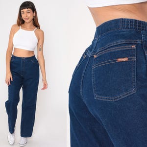 K&ouml;nnte beinhalten: Dunkelblaue Jeans mit hoher Taille und geradem Bein. Die Jeans hat ein Wrangler-Logo auf der Ges&auml;&szlig;tasche. Das Model tr&auml;gt ein wei&szlig;es Crop-Top und wei&szlig;e Turnschuhe.