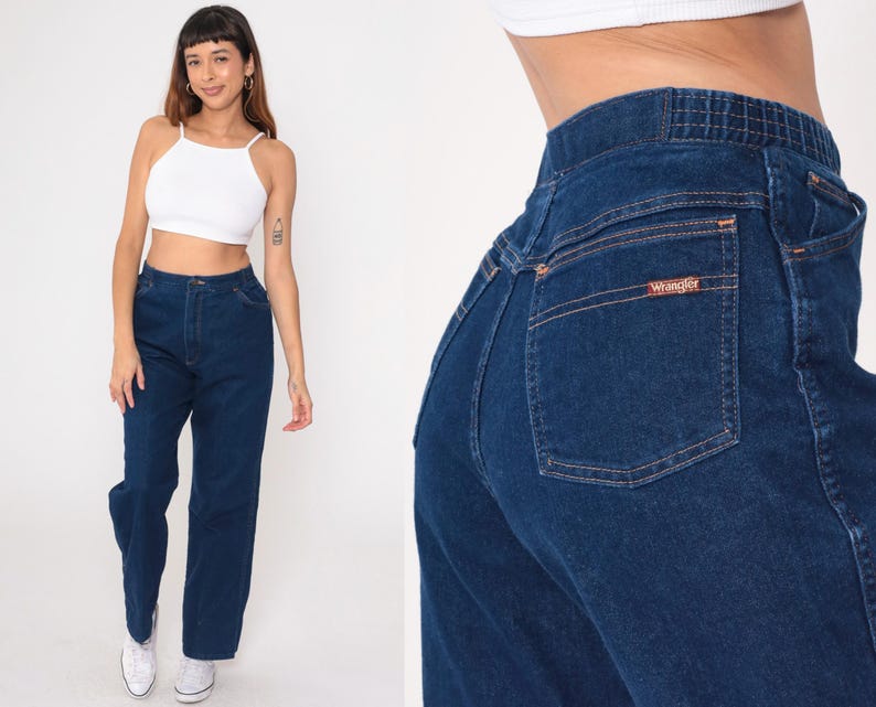K&ouml;nnte beinhalten: Dunkelblaue Jeans mit hoher Taille und geradem Bein. Die Jeans hat ein Wrangler-Logo auf der Ges&auml;&szlig;tasche. Das Model tr&auml;gt ein wei&szlig;es Crop-Top und wei&szlig;e Turnschuhe.