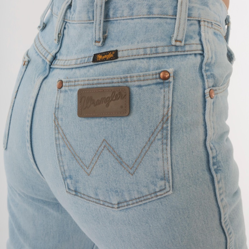 Op de afbeelding: Lichtblauwe spijkerbroek met een Wrangler-label op de achterzak. De jeans heeft een klassiek ontwerp met vijf zakken, koperen klinknagels en een bruine leren patch. Het Wrangler-logo is zichtbaar.
