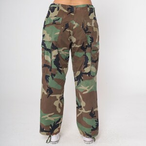 May include: Camouflage cargo pants in a woodland pattern, featuring shades of green, brown, and black. The pants have multiple pockets and a relaxed fit. The model is wearing white sneakers.