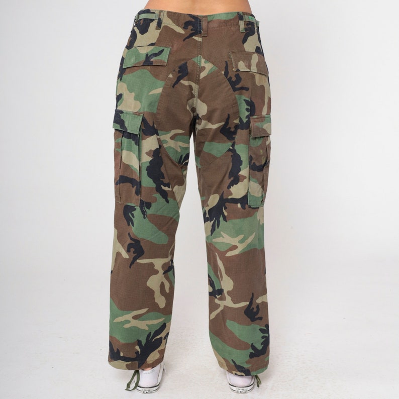 May include: Camouflage cargo pants in a woodland pattern, featuring shades of green, brown, and black. The pants have multiple pockets and a relaxed fit. The model is wearing white sneakers.