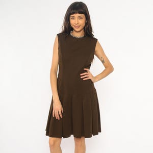Op de afbeelding: Een mouwloze, bruine jurk met een geplooide rok. De jurk heeft een eenvoudige, ronde halslijn en een getailleerd lijfje. Het model draagt een zilveren ketting. De jurk is gemaakt van een effen, donkerbruine stof.