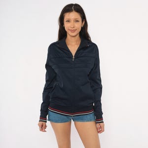 May include: A dark navy blue zip-up track jacket featuring a high collar, long sleeves, and horizontal panel detailing across the chest. The hem and sleeve cuffs are finished with thin, contrasting red and white stripes for a subtle accent.