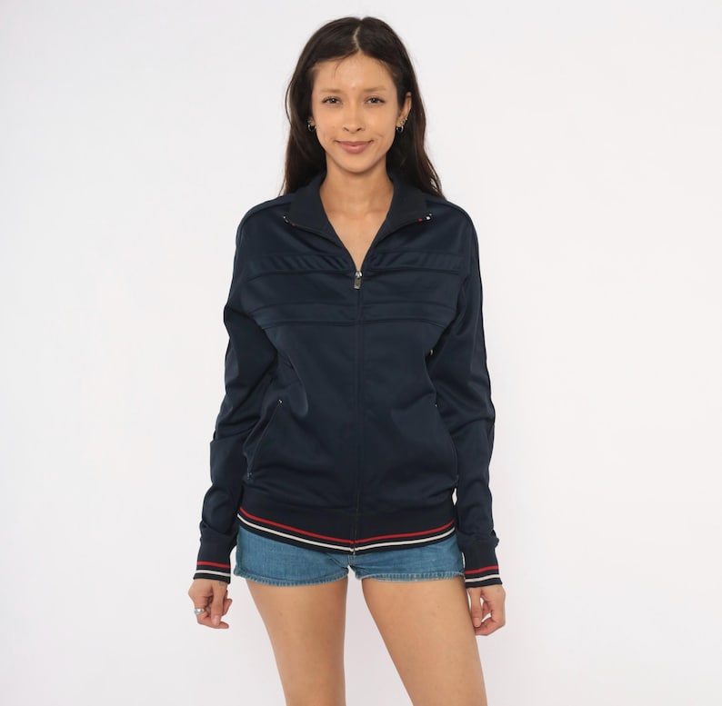 May include: A dark navy blue zip-up track jacket featuring a high collar, long sleeves, and horizontal panel detailing across the chest. The hem and sleeve cuffs are finished with thin, contrasting red and white stripes for a subtle accent.