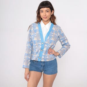 May include: A light blue cardigan with a geometric pattern in shades of blue and beige. The cardigan has a light blue trim and three gold buttons. The person is wearing a white collared shirt and denim shorts.