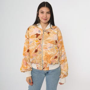 Könnte beinhalten: Gemusterte Bomberjacke mit weißem Kragen, Manschetten und Saum. Die Jacke hat ein farbenfrohes abstraktes Design in Orange-, Braun-, Rosa- und Gelbtönen. Die Jacke ist mit einem Reißverschluss versehen und wird mit einer blauen Jeans getragen.