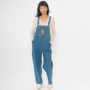 Könnte beinhalten: Blaue Jeans-Latzhose mit Winnie-the-Pooh-Stickerei auf der Latzvorderseite. Die Latzhose hat verstellbare Träger, mehrere Taschen und eine lockere Passform. Das Model trägt darunter ein cremefarbenes Langarmshirt.