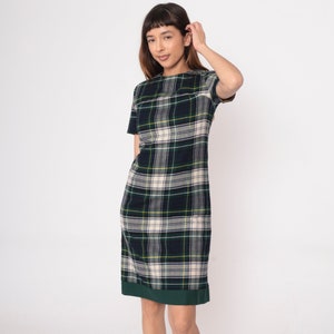 May include: A short-sleeved dress featuring a plaid pattern in shades of green, navy, and cream. The dress has a straight silhouette and a dark green hemline. The model is wearing the dress.