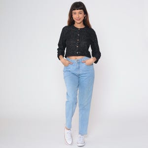 Puede incluir: Una blusa negra de manga larga con botones y un patr&oacute;n texturizado. La blusa se combina con vaqueros azul claro y zapatillas blancas. El conjunto es informal y adecuado para el uso diario.