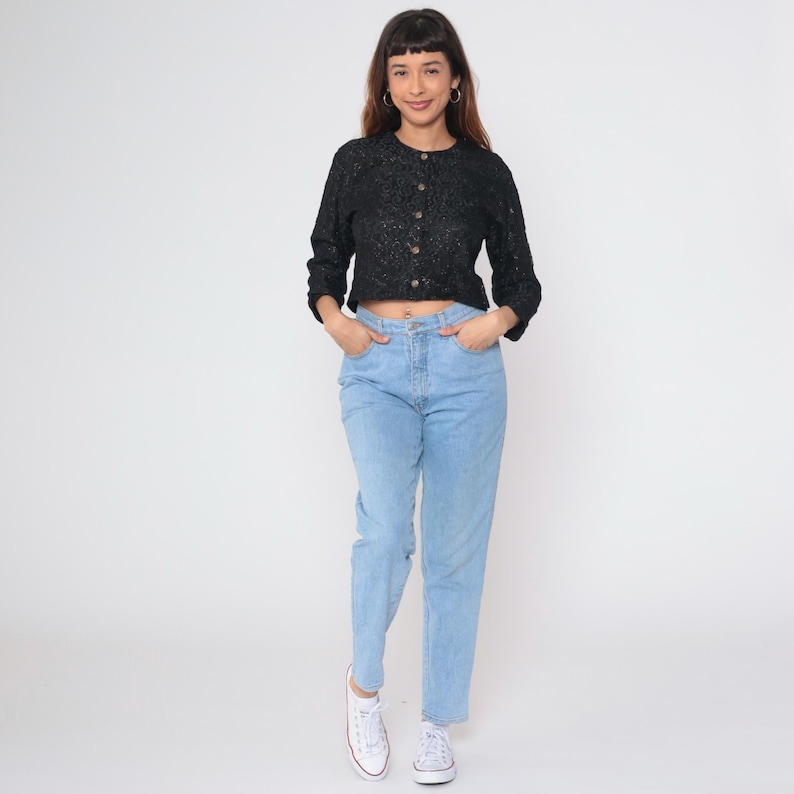 Puede incluir: Una blusa negra de manga larga con botones y un patr&oacute;n texturizado. La blusa se combina con vaqueros azul claro y zapatillas blancas. El conjunto es informal y adecuado para el uso diario.