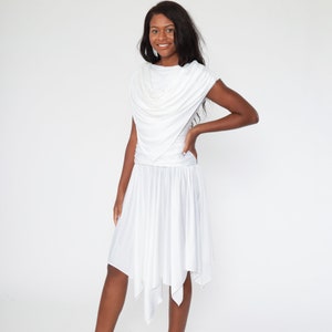 May include: A white dress with a draped cowl neckline and a flowing skirt. The dress has a fitted bodice and a flared, asymmetrical hemline. The fabric appears to be a soft, lightweight material.