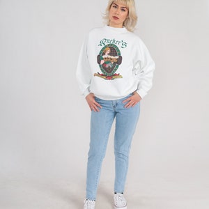 May include: White sweatshirt with a green and white logo that says "Rucker's" and features a woman holding a beer stein. The logo is surrounded by a green oval.