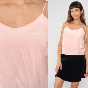 May include: A light pink camisole top with thin spaghetti straps and a V-neckline. The top is made of a silky, shiny material. The model is wearing a black skirt.
