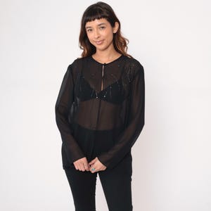 May include: A sheer black blouse with long sleeves and a button-up front. The blouse is embellished with small, sparkling details around the neckline. The model is wearing a black bra and black pants. The background is plain white.