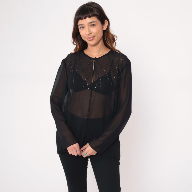 May include: A sheer black blouse with long sleeves and a button-up front. The blouse is embellished with small, sparkling details around the neckline. The model is wearing a black bra and black pants. The background is plain white.