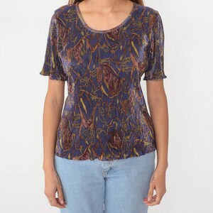 Puede incluir: Una blusa de color morado, dorado y marr&oacute;n con estampado, cuello redondo y mangas cortas. La blusa est&aacute; hecha de tela plisada.