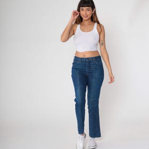 May include: A woman wearing a white tank top and blue jeans. The jeans are a classic style with a high waist and straight leg. She is also wearing white sneakers.