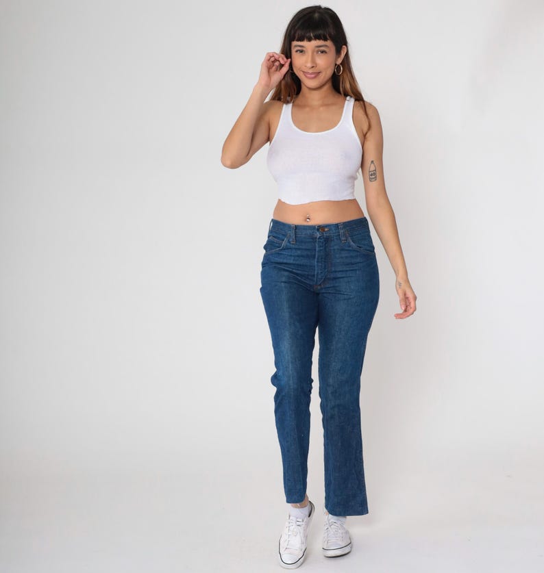 May include: A woman wearing a white tank top and blue jeans. The jeans are a classic style with a high waist and straight leg. She is also wearing white sneakers.