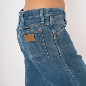 Peut inclure: Gros plan sur un jean en denim bleu avec un patch en cuir marron sur la poche arri&egrave;re. Le jean a des rivets en cuivre et une &eacute;tiquette Wrangler. Les coutures sont visibles et le jean est l&eacute;g&egrave;rement d&eacute;lav&eacute;.