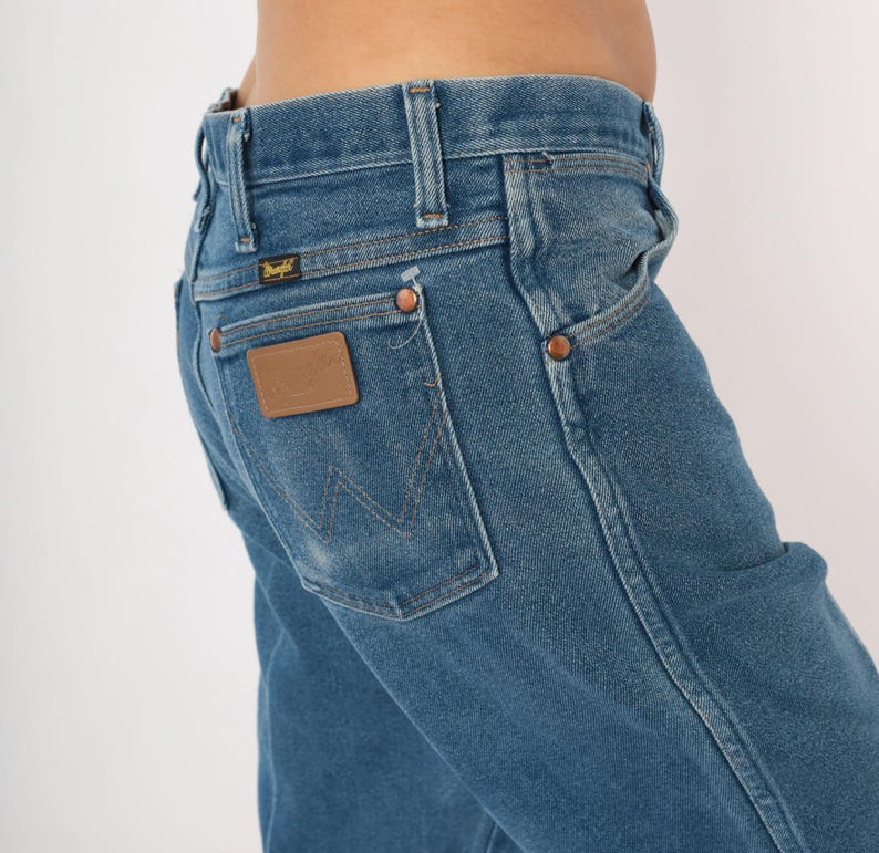 Peut inclure: Gros plan sur un jean en denim bleu avec un patch en cuir marron sur la poche arri&egrave;re. Le jean a des rivets en cuivre et une &eacute;tiquette Wrangler. Les coutures sont visibles et le jean est l&eacute;g&egrave;rement d&eacute;lav&eacute;.
