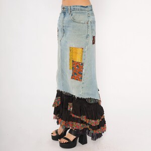 May include: A long denim skirt with patchwork details and a tiered, ruffled hem. The skirt features light blue denim with colourful fabric patches and a black ruffled bottom. The model is wearing black platform heels.