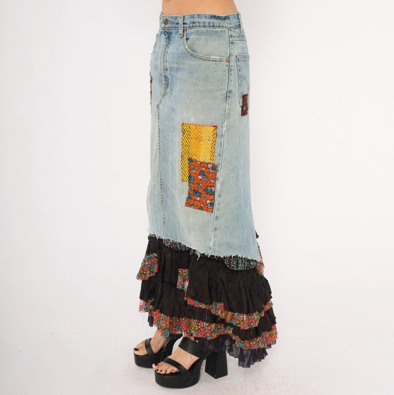 May include: A long denim skirt with patchwork details and a tiered, ruffled hem. The skirt features light blue denim with colourful fabric patches and a black ruffled bottom. The model is wearing black platform heels.