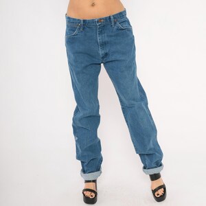 May include: Blue denim jeans with a classic five-pocket design. The jeans have a straight leg fit and are cuffed at the ankles. The jeans are worn with black platform sandals.