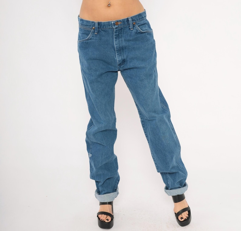 May include: Blue denim jeans with a classic five-pocket design. The jeans have a straight leg fit and are cuffed at the ankles. The jeans are worn with black platform sandals.
