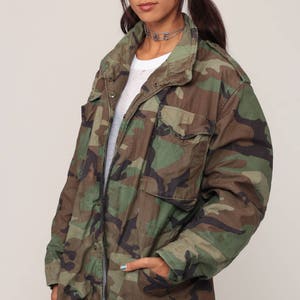 May include: A camouflage field jacket featuring a woodland pattern in shades of green, brown, and black. The jacket includes a fold-over collar, long sleeves, two chest pockets with buttoned flaps, and two lower front pockets with buttoned flaps.