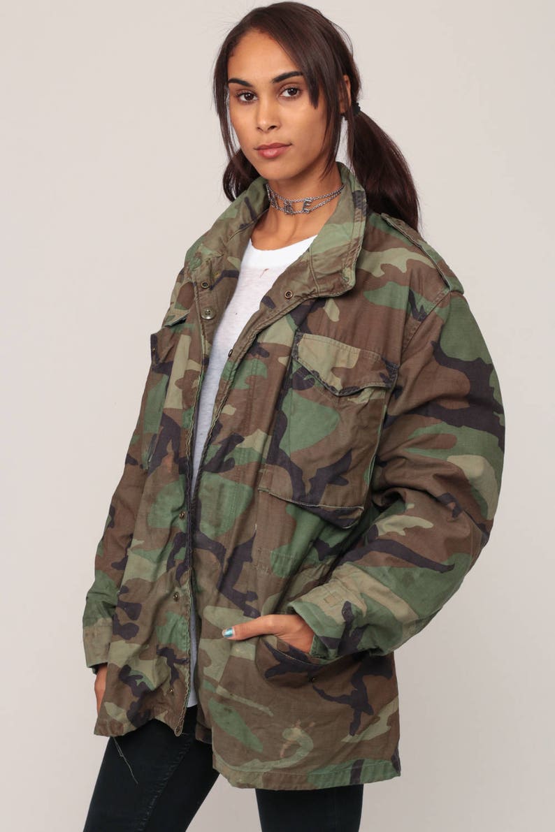 May include: A camouflage field jacket featuring a woodland pattern in shades of green, brown, and black. The jacket includes a fold-over collar, long sleeves, two chest pockets with buttoned flaps, and two lower front pockets with buttoned flaps.