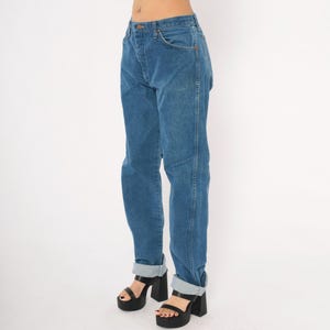 May include: Blue denim jeans with a straight leg and cuffed hem. The jeans have a classic five-pocket design and a button and zipper closure. The model is wearing black platform heels. The jeans are a medium wash.