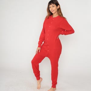 May include: A red, one-piece thermal jumpsuit with long sleeves and a button-down front. The jumpsuit is made of a textured fabric and has a relaxed fit. The model is wearing the jumpsuit. The jumpsuit is a solid red color.