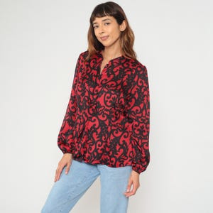 May include: A red blouse with a black ornate pattern. The long-sleeved top has a V-neck and a gathered waist. The blouse is paired with light blue jeans. The fabric appears to be a lightweight material.