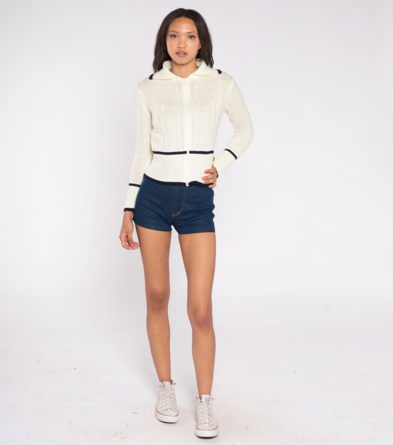 May include: A cream-colored zip-up sweater with black trim on the collar, cuffs, and waist. The sweater is paired with dark blue denim shorts and white sneakers. The outfit is displayed on a white background.