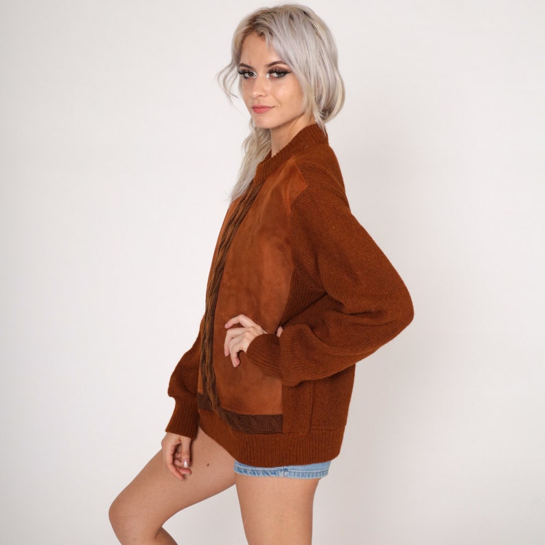 May include: A brown sweater with a suede panel on the front. The sweater has a ribbed knit texture and a relaxed fit.