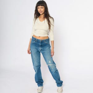 May include: A person wearing a cream-coloured, long-sleeved V-neck top and blue jeans. The jeans have a distressed patch on the right knee. The person is wearing white trainers.