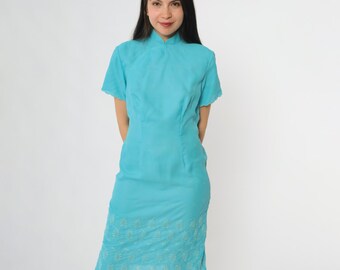 May include: A turquoise dress with a mandarin collar and short sleeves. The dress has a fitted bodice and a knee-length skirt with embroidered detailing at the hem. The dress is made of a soft, flowing fabric.