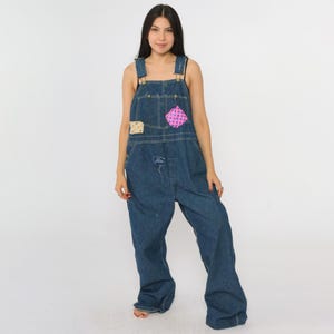 May include: A denim overall with a relaxed fit, featuring adjustable straps and multiple pockets. Patches with pink polka dots and a floral pattern add a touch of whimsy. The overall is a medium wash blue.