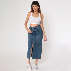 May include: A denim midi skirt with a front split, worn with a white vest top and white trainers. The skirt is a mid-wash blue and has a classic five-pocket design. The outfit is casual and stylish.