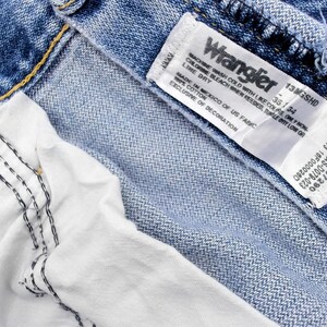 Pu&ograve; includere: Primo piano di jeans blu in denim con un'etichetta bianca Wrangler. L'etichetta mostra il nome del marchio, la taglia 35/34 e le istruzioni per la cura. I jeans hanno una fodera bianca e cuciture visibili.