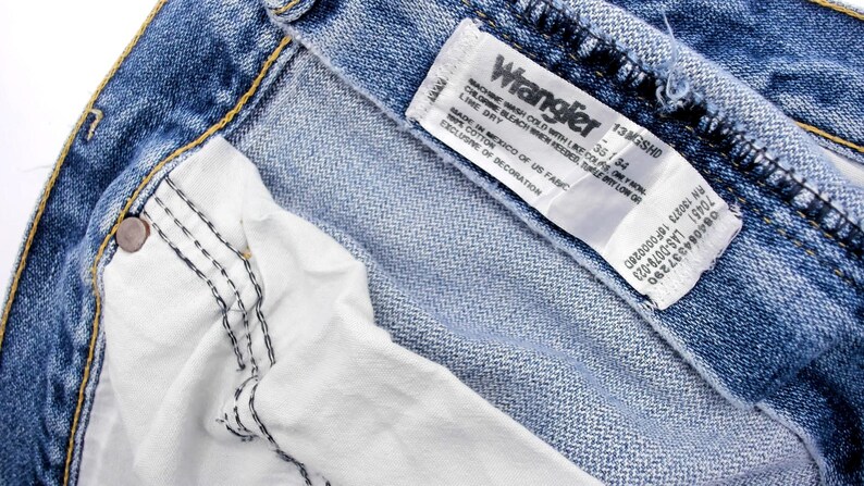 Pu&ograve; includere: Primo piano di jeans blu in denim con un'etichetta bianca Wrangler. L'etichetta mostra il nome del marchio, la taglia 35/34 e le istruzioni per la cura. I jeans hanno una fodera bianca e cuciture visibili.