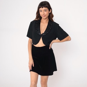 May include: Black bolero jacket with short sleeves and a satin collar adorned with small embellishments. Paired with a black velvet mini skirt. The outfit is set against a plain white background.