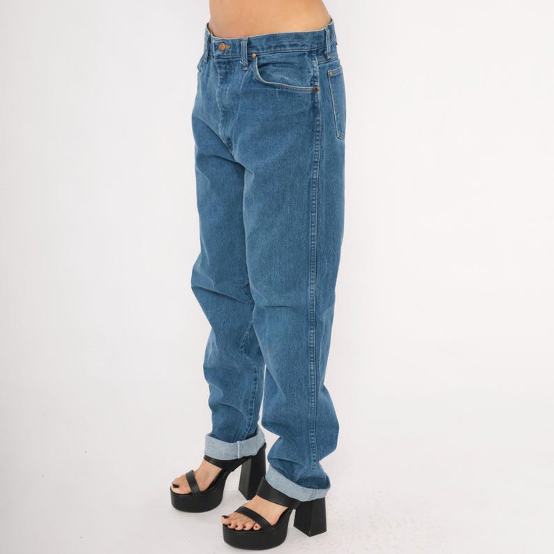 May include: Blue denim jeans with a classic five-pocket design. The jeans have a relaxed fit and are cuffed at the ankles. The model is wearing black platform heels.