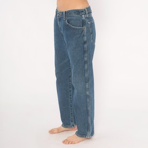 May include: Blue denim jeans with a classic five-pocket design. The jeans have a straight leg cut and a button and zipper closure. The denim is a medium wash, and the jeans appear to be worn. The person is standing barefoot.