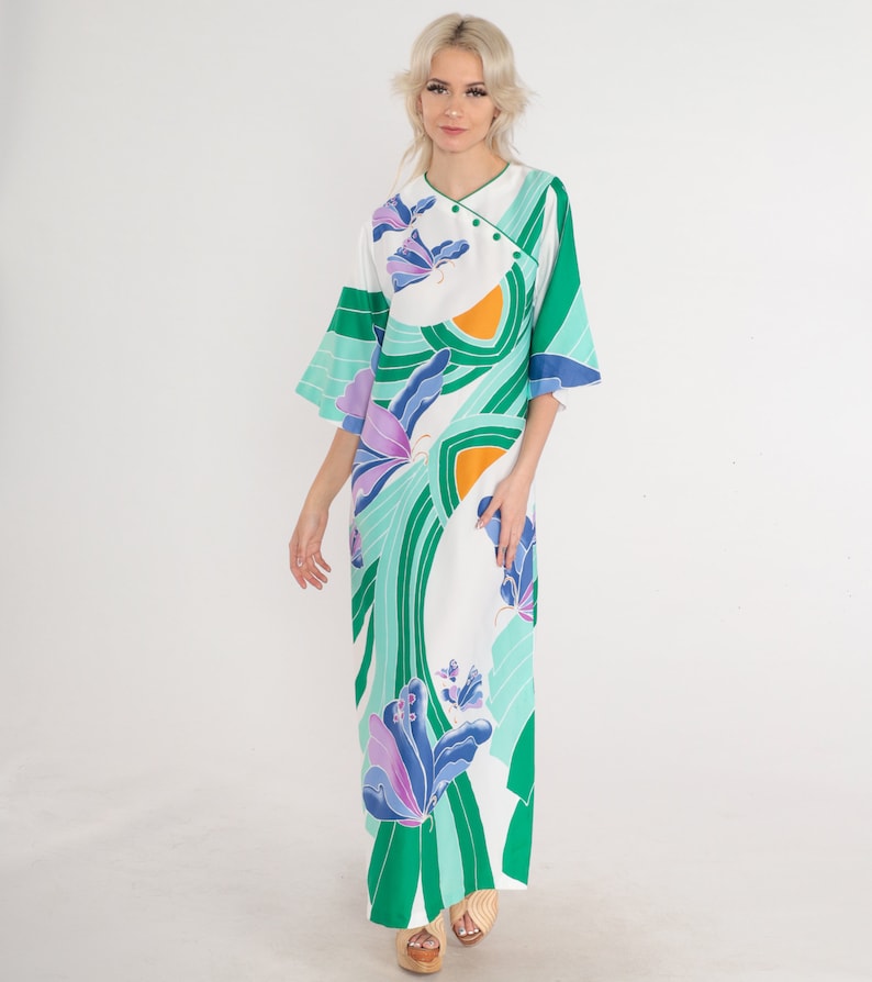 May include: A maxi dress with a white background and a vibrant green, teal, and purple abstract floral print. The dress features a mandarin collar, three-quarter sleeves, and a relaxed fit. The fabric appears to be lightweight and flowing. The dress is shown on a mannequin against a white background.