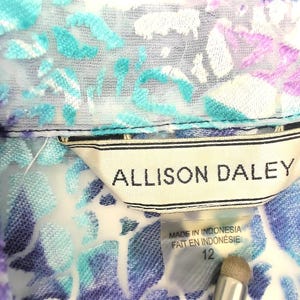 Op de afbeelding: Close-up van een blouse met bloemenpatroon en kraag. De blouse heeft een mix van blauwe, paarse en witte kleuren. Het label luidt "ALLISON DALEY" en "MADE IN INDONESIA". De maat is 12.
